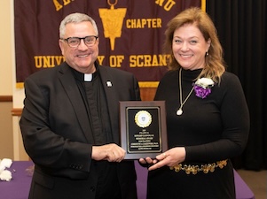A Catholic priest presents award to woman with brown hair in a black dress.