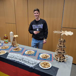 German FLTA Joshua Hartmann standing in front of a table holding a Christmas cookie. The table has a German flag on it as well as a traditional German pyramid. 