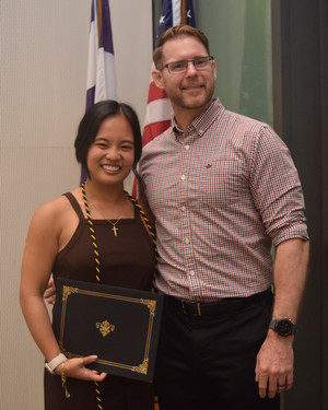 A graduate holding a diploma stands with a proud instructor in front of American flags during a graduation ceremony.