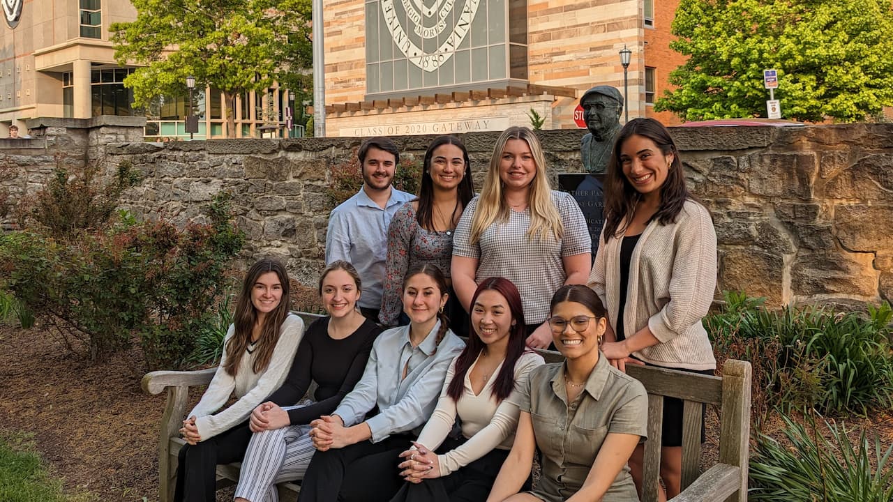 A group of nine students in the rose garden,  five sitting on a bench with four behind them.