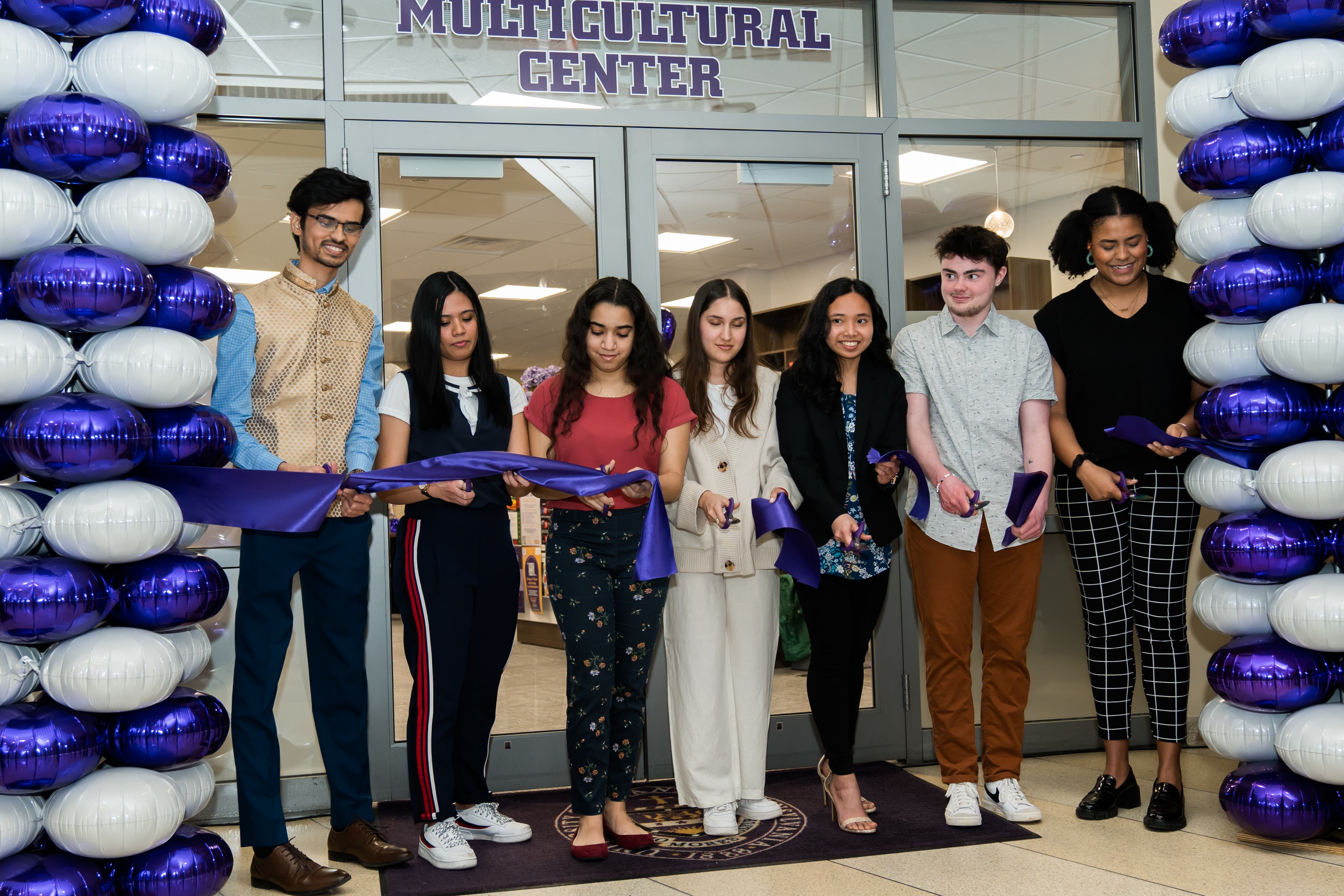 A group of seven students surrounded by purple and white balloons  cutting the ribbon at the opening of the Multicultural Center