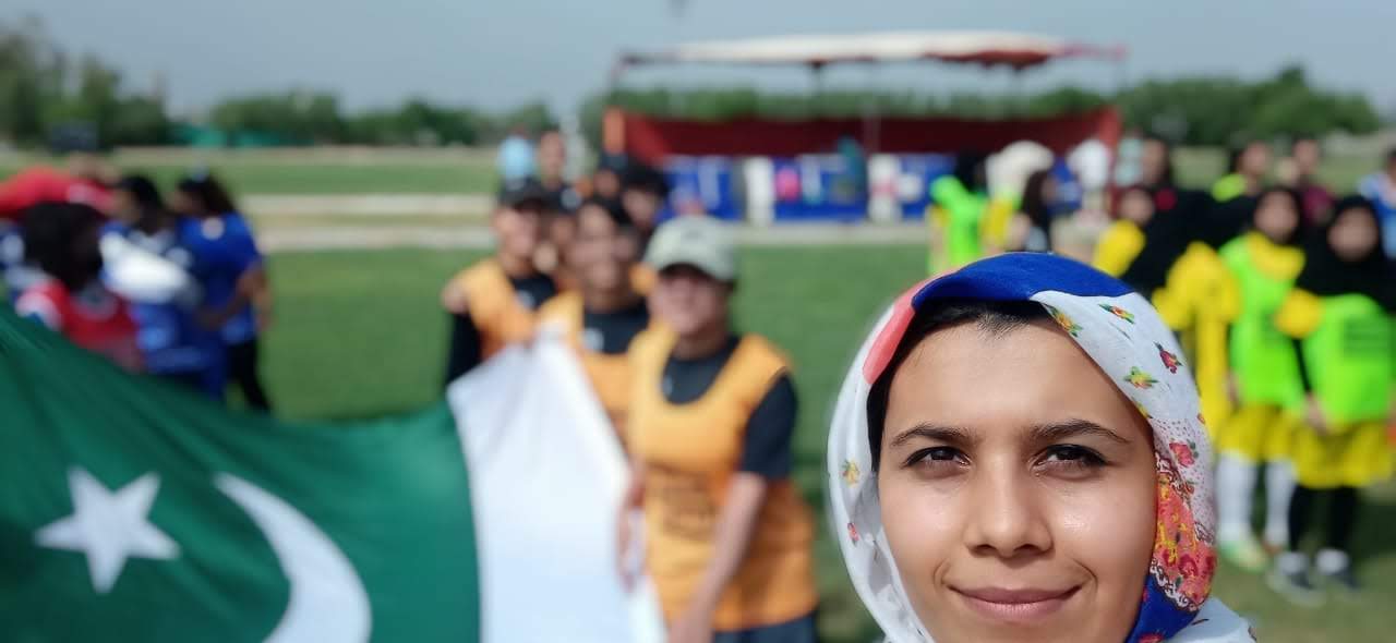 Ramsha Siddiqui in front of volunteers wearing colorful jerseys, holding the flag of Pakistan on a sports field.