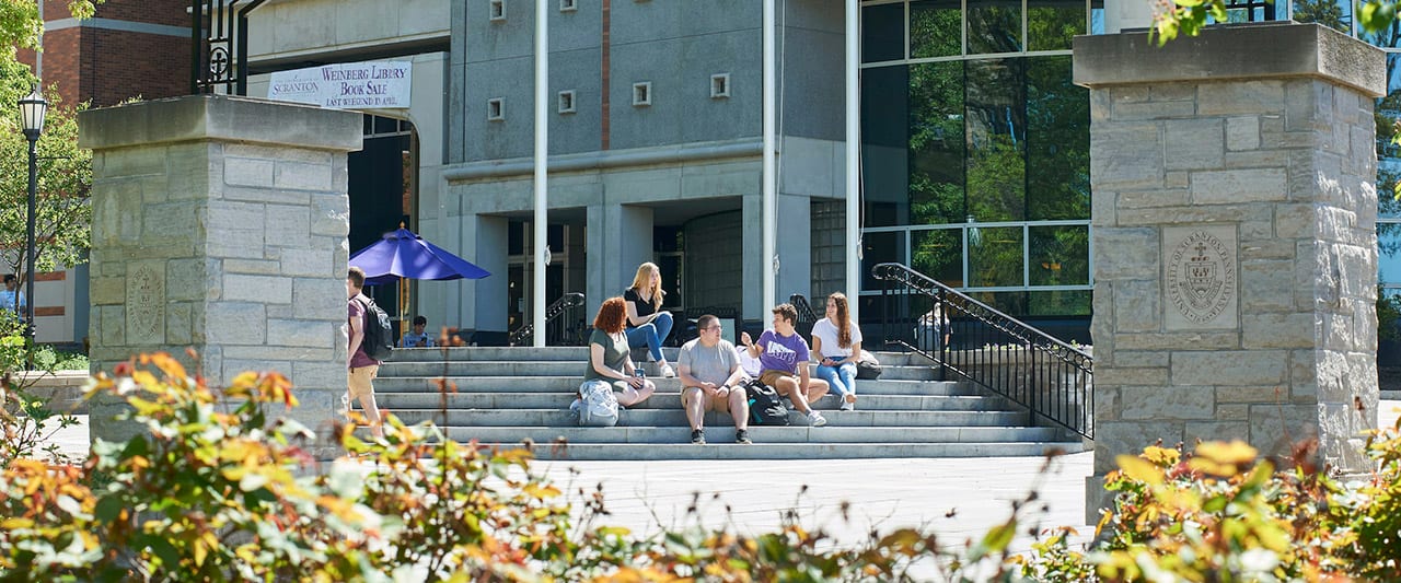 Students sitting on the steps of the Commons Flag Terrace in front of The Weinberg Memorial Library